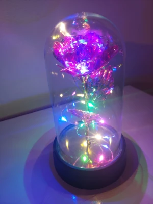 Glaskupa med LED-ros i regnbågsfärger - En dekorativ ros i metallic rosa och lila, placerad i en glaskupa med svart bas. Runt rosen slingrar sig färgglada LED-lampor som ger ett magiskt sken. Perfekt som belysning eller prydnad på skrivbordet eller hyllan. Material: glas, plast och metall.