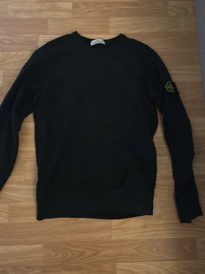 Svart sweatshirt från Stone Island - Säljer en svart sweatshirt från Stone Island med den klassiska logotypen på ärmen. Tröjan har rund halsringning och ribbade muddar vid ärmslut och nederkant. Tillverkad i mjukt bomullsmaterial som är skönt att bära.