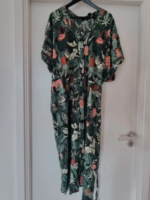 Monki Grön blommig maxiklänning kaftan med knytband  - Maxiklänning kaftan med tropiskt blommönster i grönt, vitt och brunrött. Klänningen har korta ärmar och knytband i midjan för en snygg passform. Perfekt för sommarens varma dagar och har en avslappnad, luftig stil. Knappar hela vägen så kan användas öppen. Stl XS/S men är oversized. Kan lätt användas av S-L beroende på hur man vill att den ska sitta.