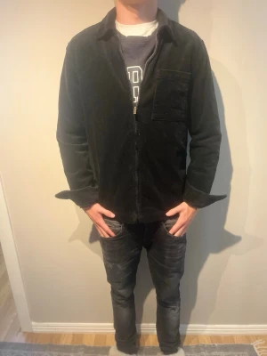 Svart overshirt i manchester - Säljer en svart overshirt i mjuk manchester med klassisk krage och bröstficka. Overshirten har raka ärmar och stängs med dragkedja framtill. Perfekt att slänga över en hoodie eller t-shirt för en avslappnad look.