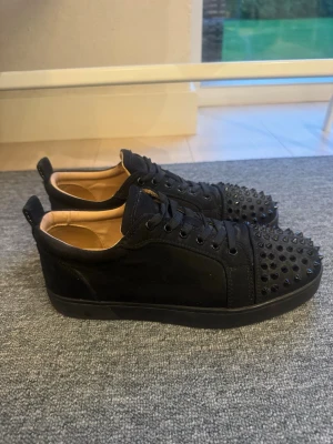 Svarta Louboutin sneakers med nitar - Säljer ett par svarta sneakers från Christian Louboutin med mattsvarta nitar på tån och klassisk röd sula. Skorna har snörning, ovandel i mocka och läderdetaljer. Perfekta för dig som vill sticka ut med en edgy och lyxig stil. Storlek 40.