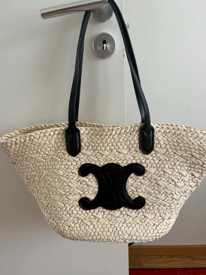 Väska  - Säljer en snygg shoppingväska  i flätad naturfärgad strå med svarta läderhandtag och stor svart -logga framtill. Perfekt för dig som vill ha en trendig och rymlig väska till alla dina grejer. Väskan har en klassisk korgform och känns riktigt lyxig.