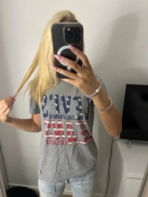 Grå Levi's t-shirt med tryck - Säljer en grå t-shirt från Levi's med stort tryck i blått och rött framtill. T-shirten har korta ärmar och klassisk passform. Materialet är mjuk bomull, perfekt till jeans eller shorts. Snygg och enkel att styla till olika outfits.