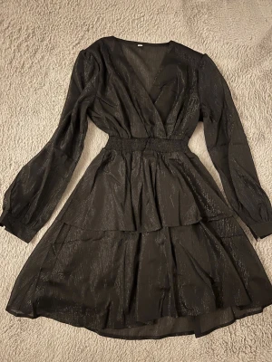 Svart omlottklänning med volanger - Snygg svart omlottklänning med v-ringning och långa ärmar. Klänningen har volangdetaljer och ett lätt glittrigt tyg som ger extra effekt. Perfekt för dig som vill ha en trendig look med markerad midja och flowy kjol. Ganska plastig