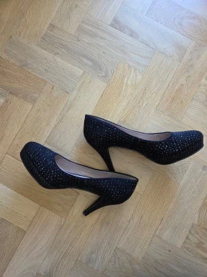 Svarta pumps med strassdetaljer från Bianco - Svarta pumps med hög klack (9 cm) och platåsula. Skorna är täckta av små glittrande strassdetaljer. Eventuellt kan några stenar saknas.