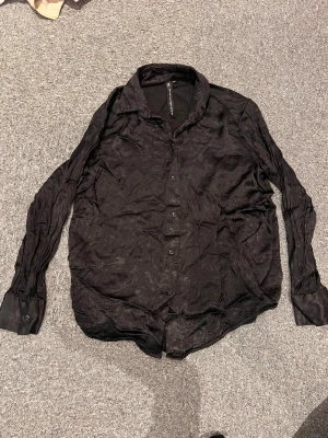 Svart skjorta från 157 Black Label XS - Säljer en svart skjorta från 157 The Black Label Collection i storlek XS. Skjortan har klassisk krage, knappar framtill och långa ärmar. Materialet har en lätt glansig finish som ger en cool look. Perfekt att styla till olika outfits.