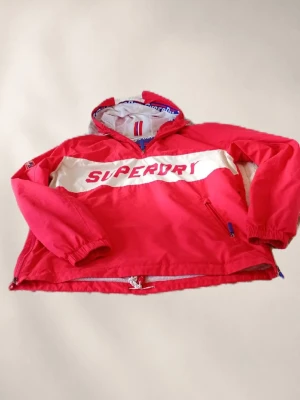 Röd vindjacka från Superdry - Cool röd vindjacka från Superdry med vit panel och stor logga framtill. Jackan har huva, dragkedja i halsen och fickor med dragkedja. Perfekt för blåsiga dagar och streetstyle. Materialet är lätt och vindtätt, med elastiska muddar och justerbar nederkant.