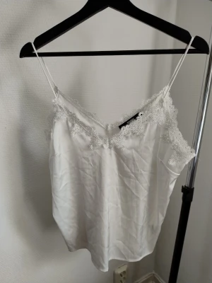 Två vita linnetoppar med spets och volang - Säljer två vita linnetoppar med tunna axelband. Ena toppen från Bik Bok har spetsdetaljer vid ringningen och den andra från Gina Tricot har en volangkant upptill. Perfekta för sommaren och lätta att styla till olika looks.