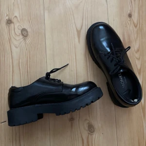 Loafers - Säljer ett par chunky svarta loafers från Monki i strl 37 med grov sula och snörning. Skorna är i blankt material och har en stilren, modern look som passar till många outfits! Defekt: Sömmen har gått upp vid hälen hos bägge skor, men inget som syns på!