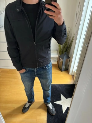 Svart vårjacka med dragkedja från Riley - Snygg svart Jacka med dragkedja framtill och två fickor med tryckknappar. Jackan har hög krage med knappdetaljer och en minimalistisk design. Perfekt för dig som vill ha en stilren och enkel look. Materialet är tunt och smidigt, vilket gör den lätt att bära.