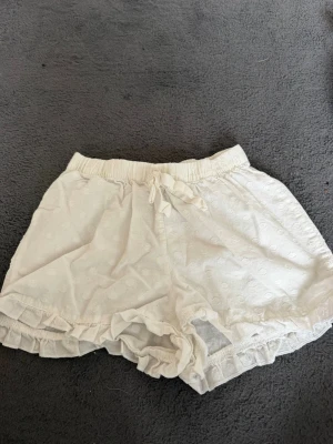 Vita shorts med volang från Primark - Säljer ett par söta vita shorts från Primark i tunn bomull med diskret prickigt mönster. Shortsen har elastisk midja med snörning och volangkant nedtill för en extra gullig touch. Perfekta för varma sommardagar!