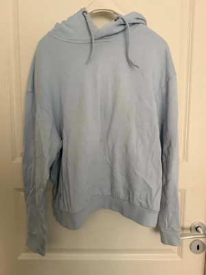 Ljusblå hoodie med huva - En enkel och stilren ljusblå hoodie med huva och dragsnören. Hoodien har långa ärmar och ribbade muddar vid ärmslut och nederkant. Finns två fläckar på framsidan av tröjan, se bild. 