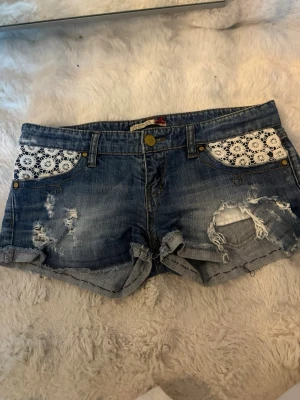 Blå jeansshorts med spetsdetaljer - Säljer ett par blå jeansshorts med slitna detaljer och råa kanter. Framfickorna har vita spetsinslag som ger en unik touch. Shortsen har klassisk femficksdesign och knappgylf. Perfekta för sommardagar och festivaler. Lågmidjwde och korta 