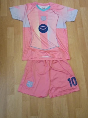 Rosa Barcelona fotbollströja och shorts - Säljer ett set med rosa och ljusblå fotbollströja och shorts från FC Barcelona. Tröjan har klubbens logga, Spotify-symbolen och mönster i pastellfärger. Shortsen har nummer 10 och klubbens emblem. Perfekt för dig som älskar fotboll och vill sticka ut på planen.