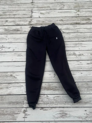 Svarta mjukisbyxor från Polo Ralph Lauren - Svarta mjukisbyxor från Polo Ralph Lauren med dragsko i midjan och resår vid bensluten. Klassisk vit logga broderad på vänster sida. Tillverkade i mjukt och bekvämt material, perfekta för chill dagar eller när du vill ha en avslappnad stil.
