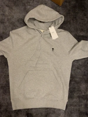 Ami Paris hoodie - Snygg grå hoodie från Ami Paris med klassisk huva och dragsko. Framtill finns en liten svart Ami-logga broderad på bröstet och en stor magficka. Tillverkad i mjuk bomull för skön känsla och avslappnad stil.