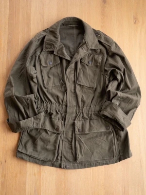 Vintage Military Olivgrön Field Jacket, size S - Snygg olivgrön militär parkajacka med fyra stora fickor framtill och justerbar midja. Jackan har klassisk krage, långa ärmar och knäppning med både knappar och dragkedja. Perfekt för lager på lager och streetwear-vibe.