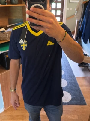 Blå officel Sverige fotbollströja Adidas - Mörkblå fotbollströja från Adidas med gula detaljer och Sveriges landslagsemblem på bröstet. Tröjan har korta ärmar, V-ringad krage och gula ränder på axlarna. Perfekt för match eller träning.