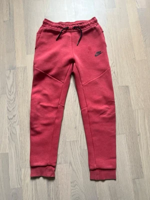 Röda Nike Tech Fleece mjukisbyxor - Säljer ett par röda Nike Tech Fleece mjukisbyxor med svarta detaljer och dragsko i midjan. Byxorna har ribbade muddar vid bensluten, fickor på sidorna och en extra ficka med dragkedja bak. Perfekta för en chill och sportig look.