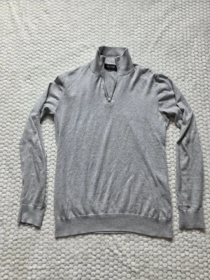 1/4 zip tröja från Dressmann - Ljusgrå tröja från Dressmann med 1/4 dragkedja och hög krage. Tillverkad i mjuk bomull, perfekt för lager på lager. Långärmad modell med ribbade muddar vid ärmslut och nederkant. Enkel och stilren design som passar till det mesta. Priset kan självklart diskuteras!