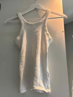 Vit ribbad linnetopp - Säljer en enkel och stilren vit ribbad linnetopp med breda axelband och rundad halsringning. Perfekt att matcha med jeans eller kjol för en clean look. Materialet är mjukt och stretchigt för skön passform.