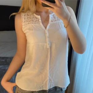 Vit ärmlös blus med spets från Soaked:  - Supersöt vit blus från Soaked med ärmlös design och spetsdetaljer upptill. Blusen har knappar framtill och ett luftigt, lätt material som passar perfekt till jeans eller kjol. Spetsen har rutigt mönster och ger en romantisk vibe. Perfekt för varma dagar!