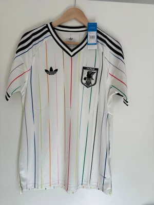 Adidas Japan vit fotbollströja M - Snygg vit fotbollströja från Adidas med Japan-emblem på bröstet och tunna färgglada ränder i blått, rött, grönt och gult. Svarta detaljer på axlar och krage. Tillverkad i lätt och ventilerande material, perfekt för träning eller match.