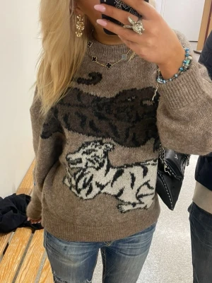Brun stickad tröja  - Cool brun stickad tröja med stor svart och vit tiger på framsidan. Tröjan har avslappnad passform, rund halsringning och ribbade muddar vid ärmar och nederkant. Perfekt för dig som gillar djurmönster och vill sticka ut lite extra.