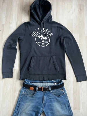 Svart hoodie från Hollister - Svart hoodie från Hollister med vit logga och palmtryck på bröstet. Klassisk känguruficka framtill och justerbar huva med snören. Tillverkad i mjukt bomullsmaterial som är skönt att ha på sig. Perfekt för en avslappnad och cool stil. Storlek S