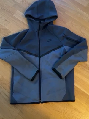 Nike Tech Fleece hoodie - Nike Tech Fleece hoodie i två nyanser av grått med hel dragkedja och huva. Snygg design med svarta detaljer och diskret Nike-logga på bröstet. Materialet är mjukt och lätt, perfekt för en avslappnad och sportig look.