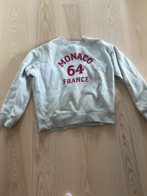 Grå sweatshirt med Monaco-tryck - Säljer en grå sweatshirt med rött tryck på ryggen där det står 'MONACO 64 FRANCE'. Tröjan har rund halsringning, ribbade muddar vid ärmslut och nederkant. Perfekt för dig som gillar sportig och avslappnad stil.
