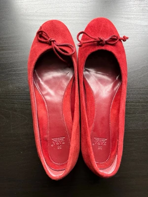 Röda mocka ballerinaskor från Zara - Snygga röda ballerinaskor från Zara i mjuk mocka med rund tå och dekorativ rosett framtill. Skorna har en klassisk siluett och helröd insida för en stilren look. Perfekta för dig som vill ha en färgklick till outfiten. Dom är str 38, men dom är  lita småa i storleken, så jag skulle säga de är närmare en 37