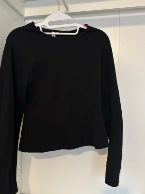 Svart långärmad topp - En enkel svart långärmad topp med rund halsringning. Modellen är rak och något croppad, vilket ger en modern och stilren look. Perfekt att matcha med jeans eller kjol för en clean outfit.