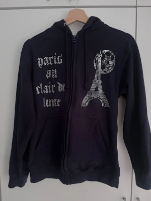Mörkblå hoodie från bling lucid  - Cool mörkblå hoodie med dragkedja och luva. Framsidan har strasstenar som bildar texten 'Paris au clair de lune' samt Eiffeltornet och måne i strass. Mjuk insida och känguruficka framtill. Perfekt för dig som gillar statement-plagg och Paris-vibes.