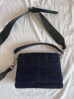 Mörkblå axelväska i tweed - Snygg mörkblå axelväska i tweed med bred svart axelrem i läderimitation och silverfärgade metalldetaljer. Väskan har en rektangulär form och lockstängning. Perfekt för dig som vill ha en trendig och unik accessoar till din outfit.