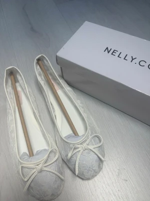 Vita spetsballerinaskor från Nelly - Säljer ett par vita ballerinaskor från Nelly med elegant spetsmaterial och dekorativ rosett framtill. 💕Helt nya och oanvända💗 slutsålda på hemsidan!!😊