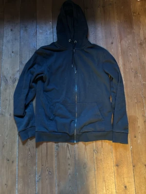 Svart hoodie med dragkedja - Klassisk svart hoodie med huva och dragsko, hel dragkedja framtill och två stora fickor. Perfekt för en avslappnad stil och enkel att matcha med det mesta. Tillverkad i mjukt material för extra komfort.