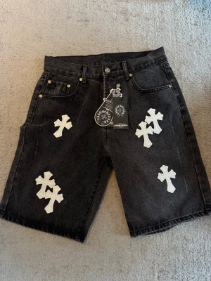 Svarta Chrome Hearts shorts - Svarta denimshorts från Chrome Hearts med vita korsapplikationer i skinn. Klassisk femficksmodell med knappgylf och detaljer i silver. Snygg streetwear-look med unika detaljer som sticker ut.