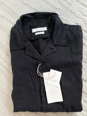 Svart linneskjorta från Zara - Stilren svart skjorta från Zara i 100% linne. Skjortan har korta ärmar, klassisk krage och knäppning framtill. Perfekt för en avslappnad men snygg look. Enkel att matcha med både jeans och shorts. Nypris 499kr