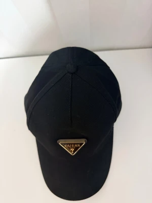 Svart keps från Guess med logga - Snygg svart keps från Guess med den ikoniska trekantiga metalloggan framtill i guld och svart. Klassisk böjd skärm och justerbar passform bak. Tillverkad i slitstark bomull som passar perfekt till streetwear och casual outfits.