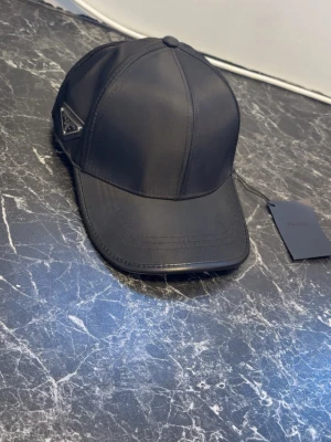 Svart keps från Prada i nylon - Snygg svart keps från Prada i glansigt nylonmaterial. Klassisk böjd skärm och sexpanelers design med diskret Prada-logga på sidan. Perfekt accessoar för en clean och modern look.