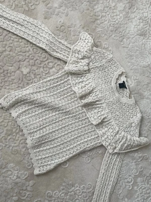 Vit stickad topp med volang - Supersöt vit stickad topp med volangdetaljer längs axlarna och ett fint hålmönster. Tröjan har långa ärmar och är gjord i ett mjukt, tunt material som känns luftigt. Perfekt för dig som gillar romantisk och trendig stil.