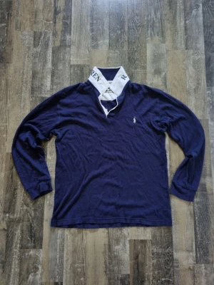 Marinblå långärmad skjorta Ralph Lauren - Snygg marinblå långärmad skjorta från Ralph Lauren med vit krage där märkesnamnet står tryckt. Klassisk passform med liten broderad logga på bröstet. Perfekt för dig som gillar stilrena plagg med en twist.