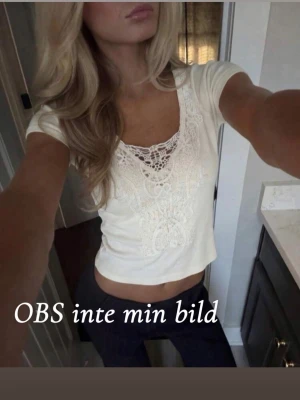 Vit  topp med spetsdetaljer - Säljer en ljusvit croppad topp med snygga spetsdetaljer framtill. Toppen har korta ärmar och en v-ringad halsringning med dekorativ spets som ger en romantisk vibe. Perfekt att styla med jeans eller kjol för en trendig look.