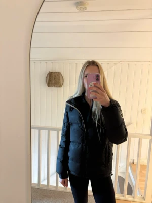 Svart pufferjacka från D brand - Säljer en svart pufferjacka från D brand med broderad logga på ärmen. Jackan har hög krage, dragkedja framtill och flera fickor. Perfekt för kalla dagar och riktigt skön att bära. Klassisk modell som passar till det mesta.