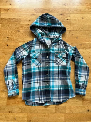 Rutig hoodie-skjorta från Hollister - Snygg rutig skjorta från Hollister med huva och knappar framtill. Skjortan har blå, turkosa, vita och grå toner, två bröstfickor med knapp och långa ärmar. Perfekt för en avslappnad streetstyle-look.