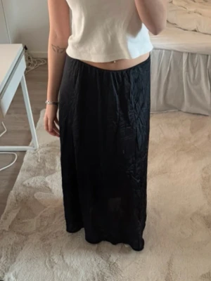 Svart långkjol i satin-look - Säljer en svart långkjol med snygg satin-look och lätt glansig yta. Kjolen har en rak passform och faller mjukt längs benen. Perfekt att styla med crop tops eller stickade tröjor för en trendig vibe.