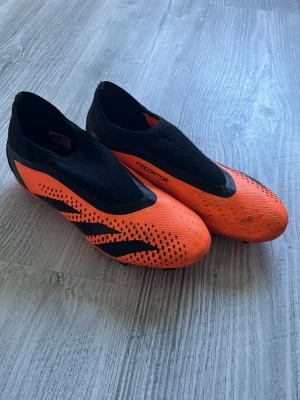 Adidas Predator orange fotbollsskor - Säljer ett par grymma Adidas Predator fotbollsskor i orange och svart. Skorna har en strömlinjeformad design utan snörning, med mönstrade detaljer och klassiska tre svarta ränder på sidan. Ovandelen är i syntetmaterial och skon är byggd för bra grepp och kontroll på planen.