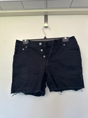 Svarta jeansshorts med rå kant W28 Weekday - Snygga svarta jeansshorts med hög midja och rå, fransig kant nedtill. Shortsen har knappgylf med fyra silverfärgade knappar och klassiska fem fickor. Perfekta för en avslappnad och trendig sommarlook.