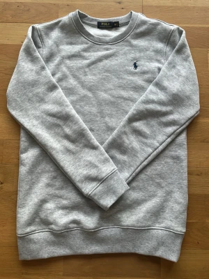 Grå sweatshirt från Polo Ralph Lauren - Klassisk grå sweatshirt från Polo Ralph Lauren med rund halsringning och diskret blå logga broderad på bröstet. Tröjan har ribbade muddar vid ärmslut och nederkant. Perfekt basic-plagg i mjukt material.